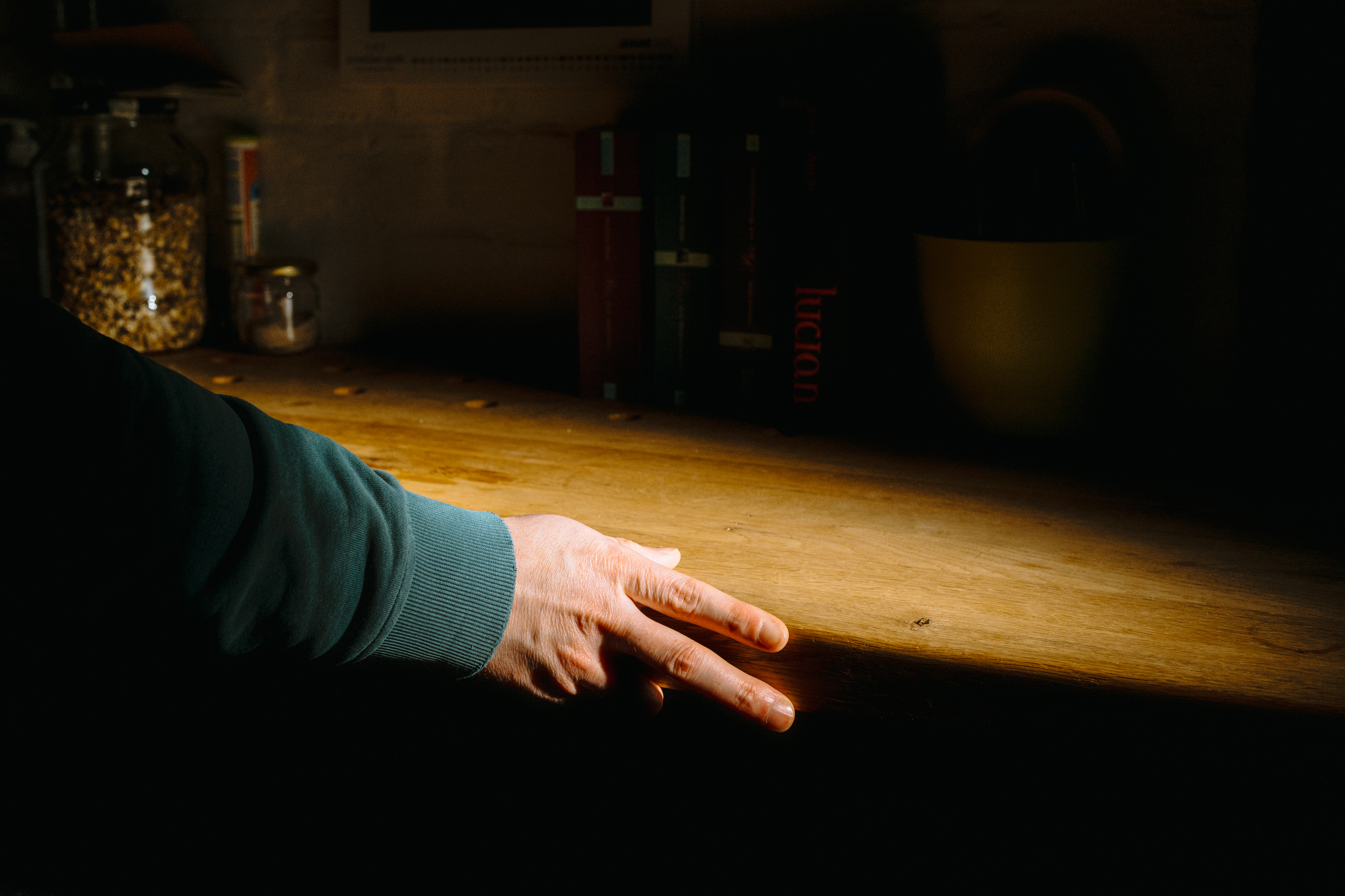 Detailaufnahme in einer dunklen Umgebung: Eine Hand ruht auf einer hölzernen Arbeitsplatte, die von einem warmen, punktuellen Lichtstrahl beleuchtet wird. Im Hintergrund sind schemenhaft Vorratsgläser und Bücherrücken zu erkennen.