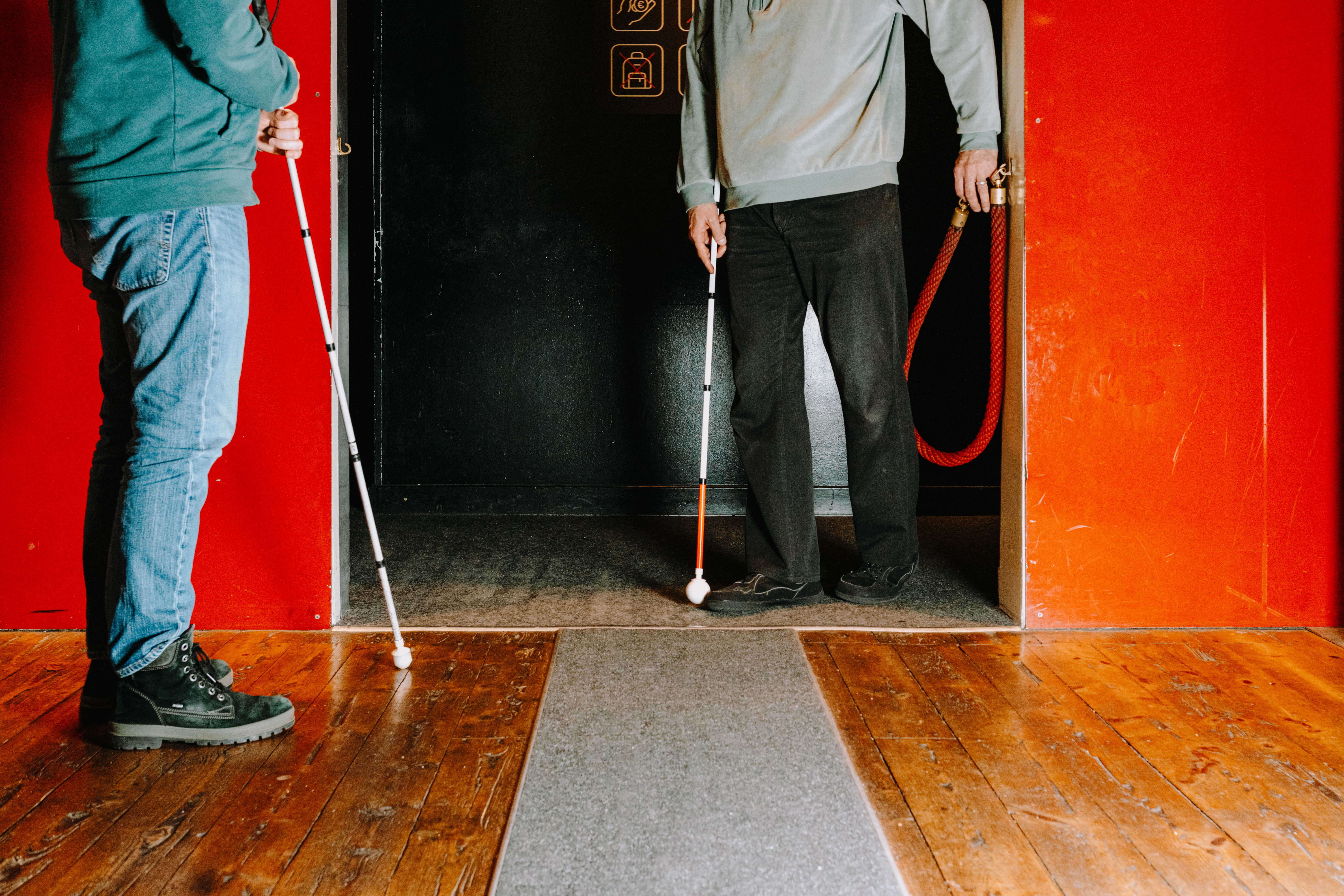 Die Aufnahme zeigt den unteren Körperabschnitt zweier Personen in einem Innenraum mit Holzdielenboden. Beide Personen halten einen weißen Langstock für blinde und sehbehinderte Menschen in der Hand. Eine Person im linken Vordergrund trägt Jeans und dunkle Schnürstiefel, während die zweite Person im Hintergrund an einer Türöffnung mit roten Wänden steht. Zwischen ihnen verläuft ein grauer Teppichläufer auf dem Boden.