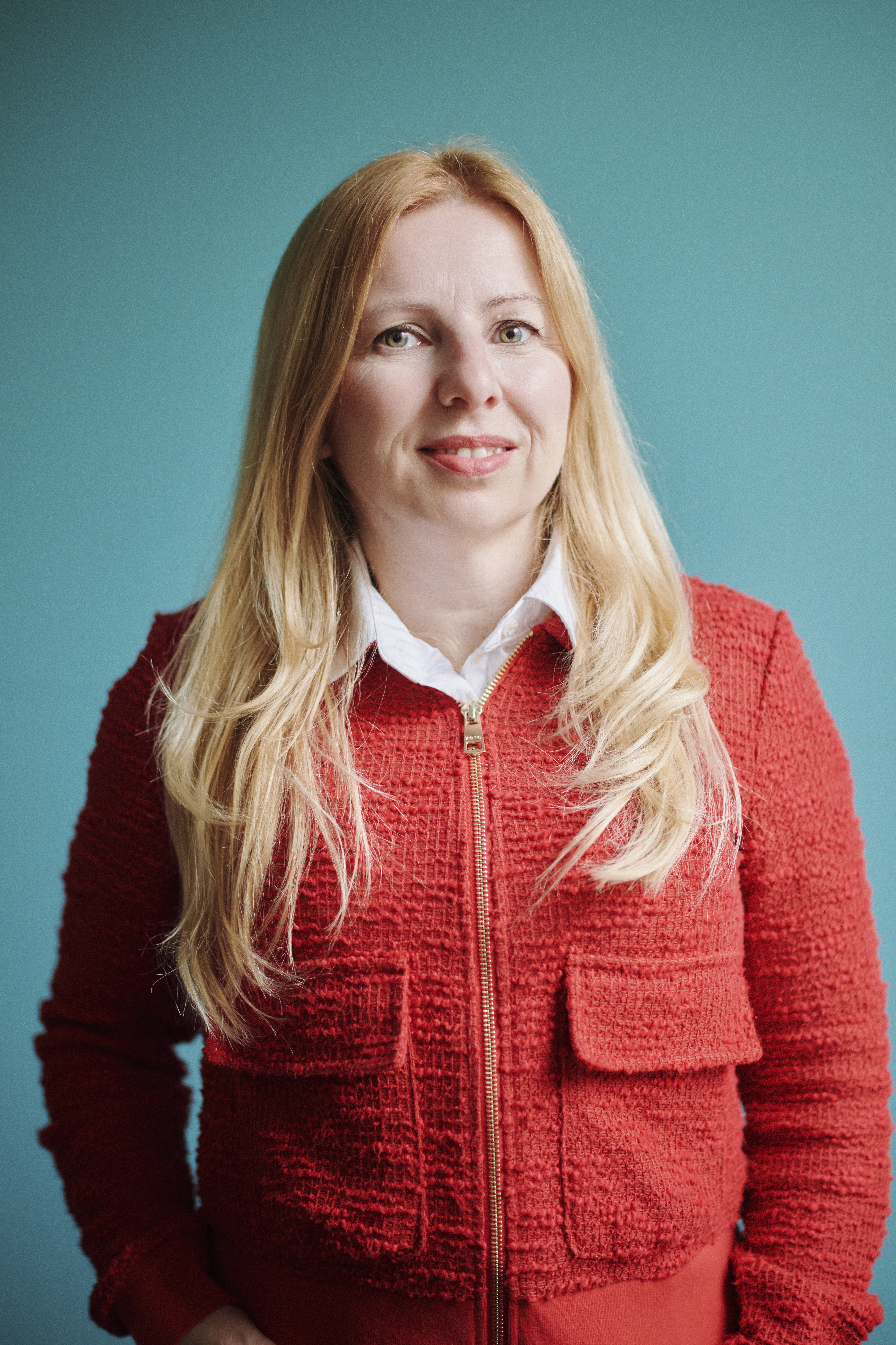 Porträtaufnahme einer lächelnden Frau mit langen blonden Haaren vor einem türkisfarbenen Hintergrund. Sie trägt eine rote Strickjacke mit Reißverschluss über einer weißen Bluse.