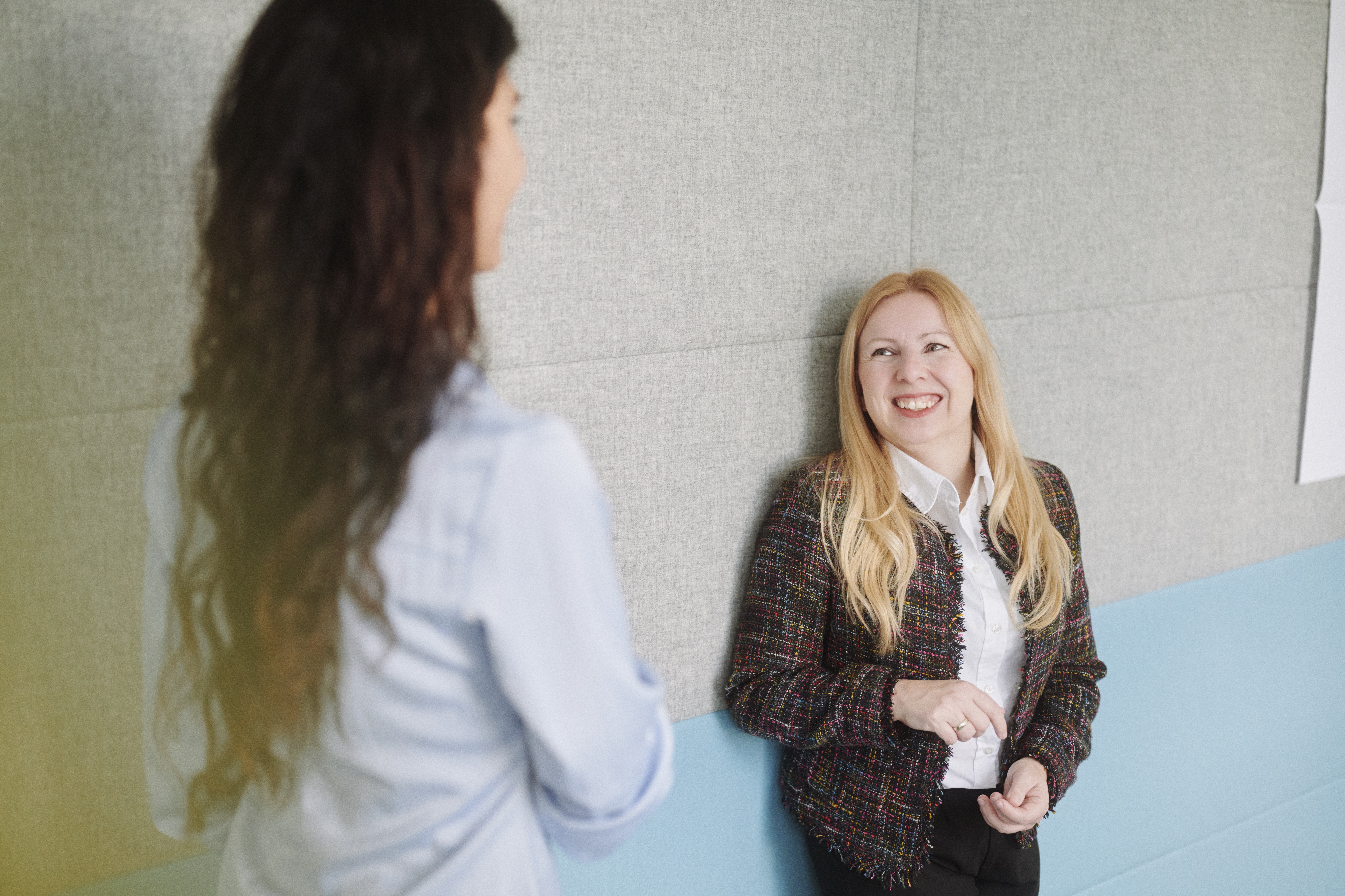 Die blondhaarige Frau in der Tweed-Jacke lehnt lachend an einer Bürowand und unterhält sich mit einer Kollegin mit langen dunklen Locken, die im Vordergrund mit dem Rücken zur Kamera steht.