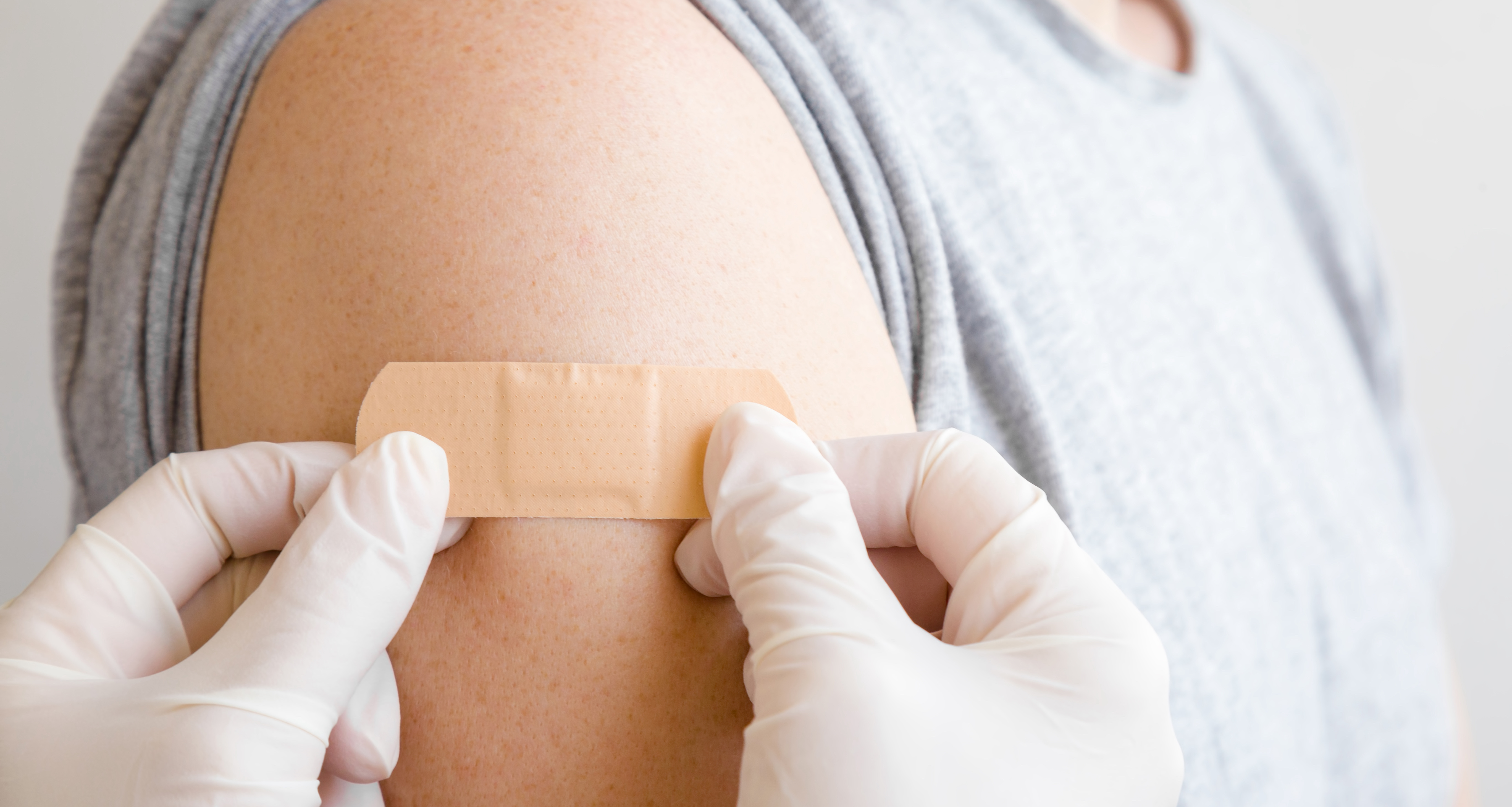 Foto: Ein Plaster wird auf einen entblößten Oberarm geklebt. Die Person, die das Pflaster aufklebt, trägt weiße Laborhandschuhe.