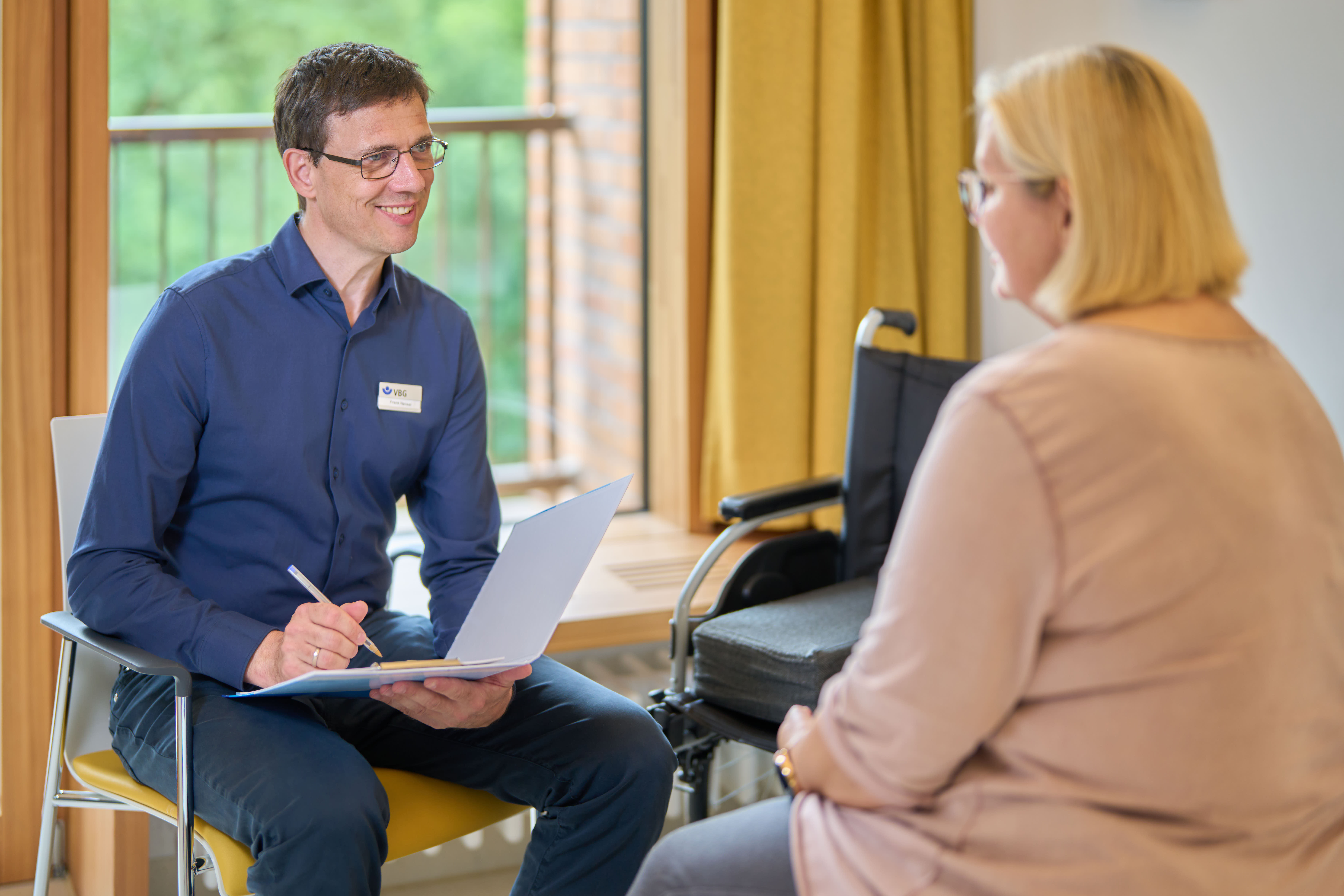 Foto: Frank Hensel sitzt im Patientenzimmer einer Rehabilitandin gegenüber. Er lächelt freundlich und macht sich Notizen auf seinem Klemmbrett.