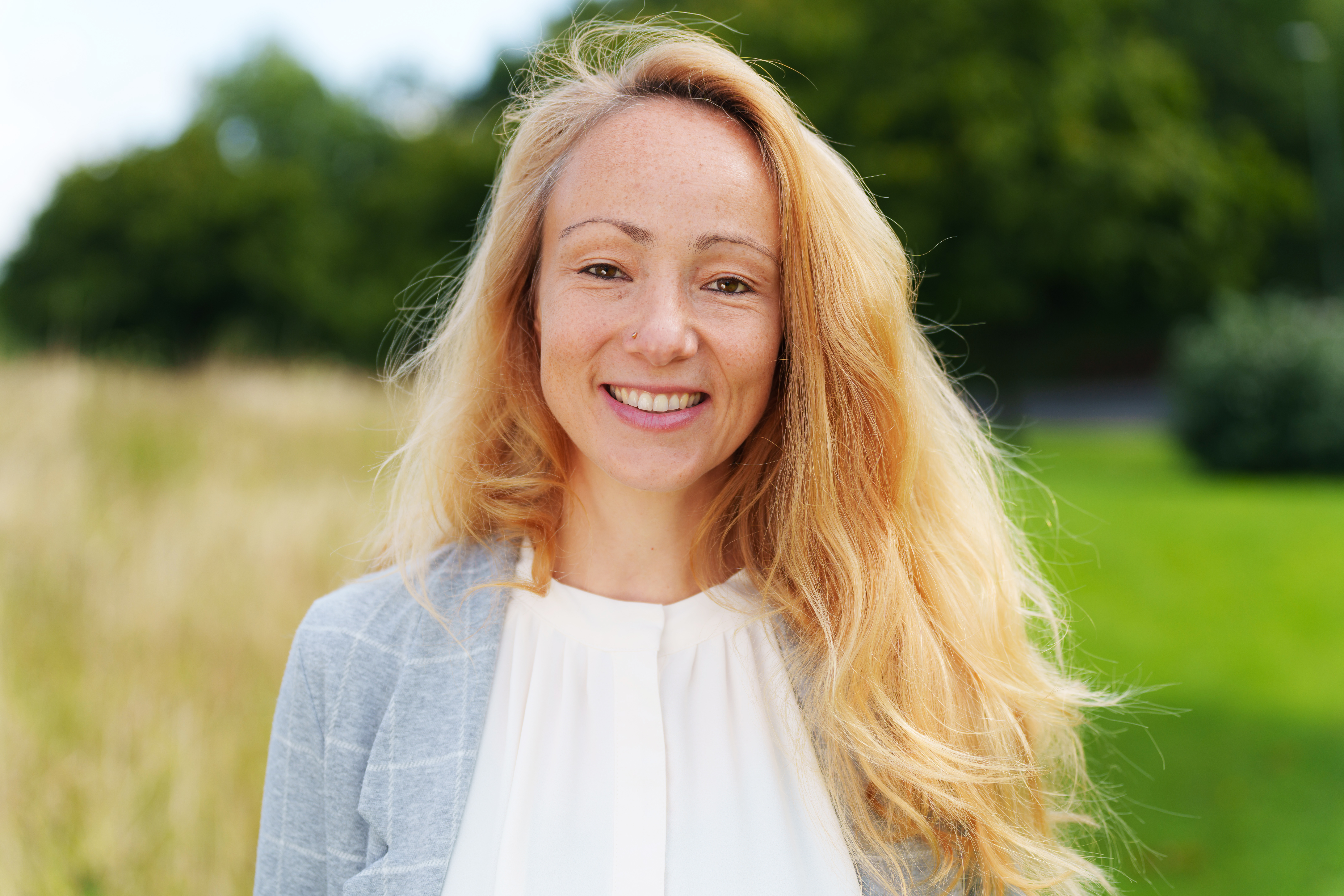      Eine lächelnde Frau mit langen blonden Haaren steht im Freien vor einem unscharfen Hintergrund aus Bäumen und Wiesen. Sie trägt eine helle Bluse und einen grauen Blazer.