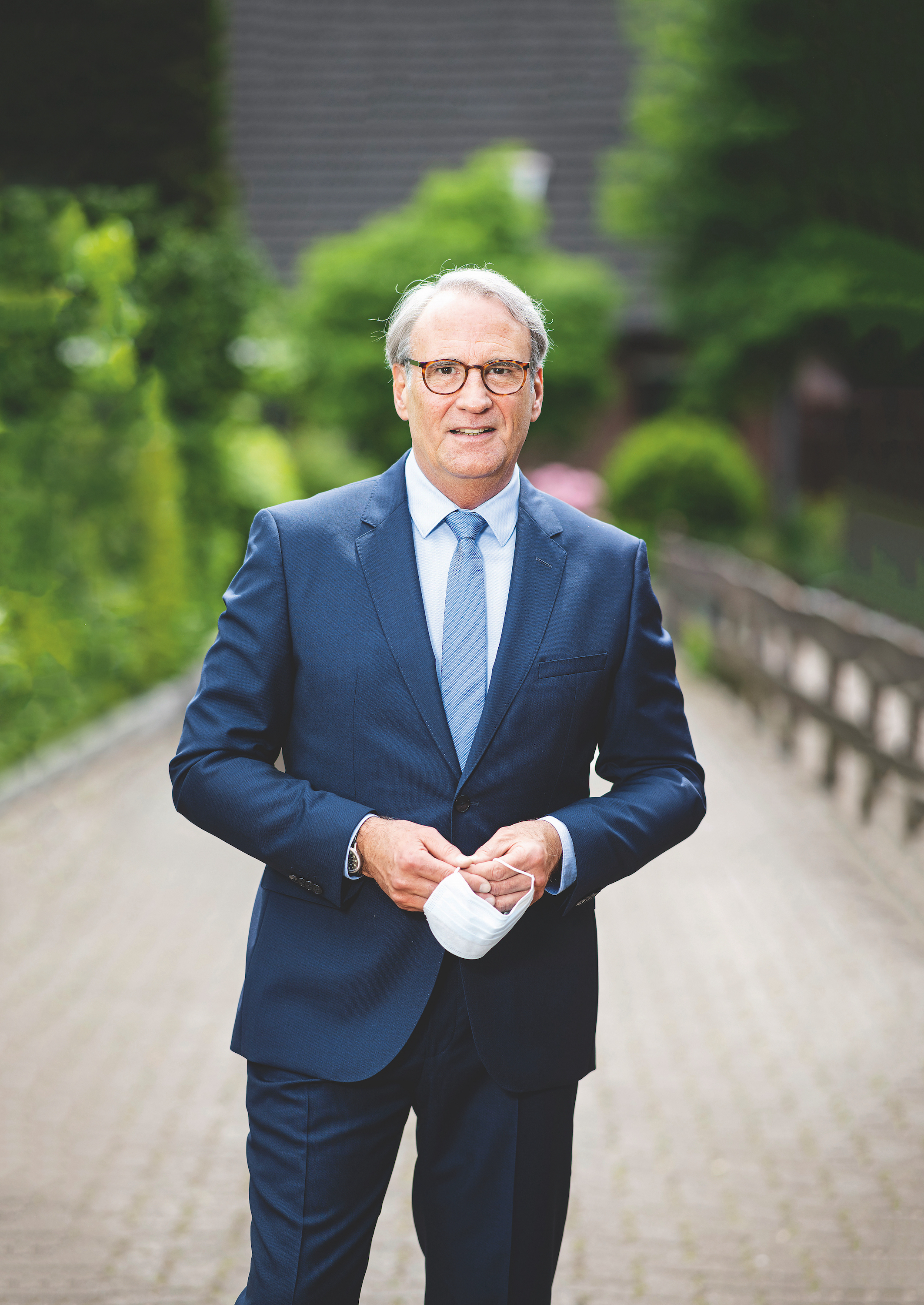 Dr. Jens Petersen leitet den Fachbereich Arbeitsmedizin und Fürsorge bei der VBG. Foto: Dr. Jens Petersen steht im Freien und schaut in die Kamera. Er trägt einen Anzug und hält eine Maske in den Händen.