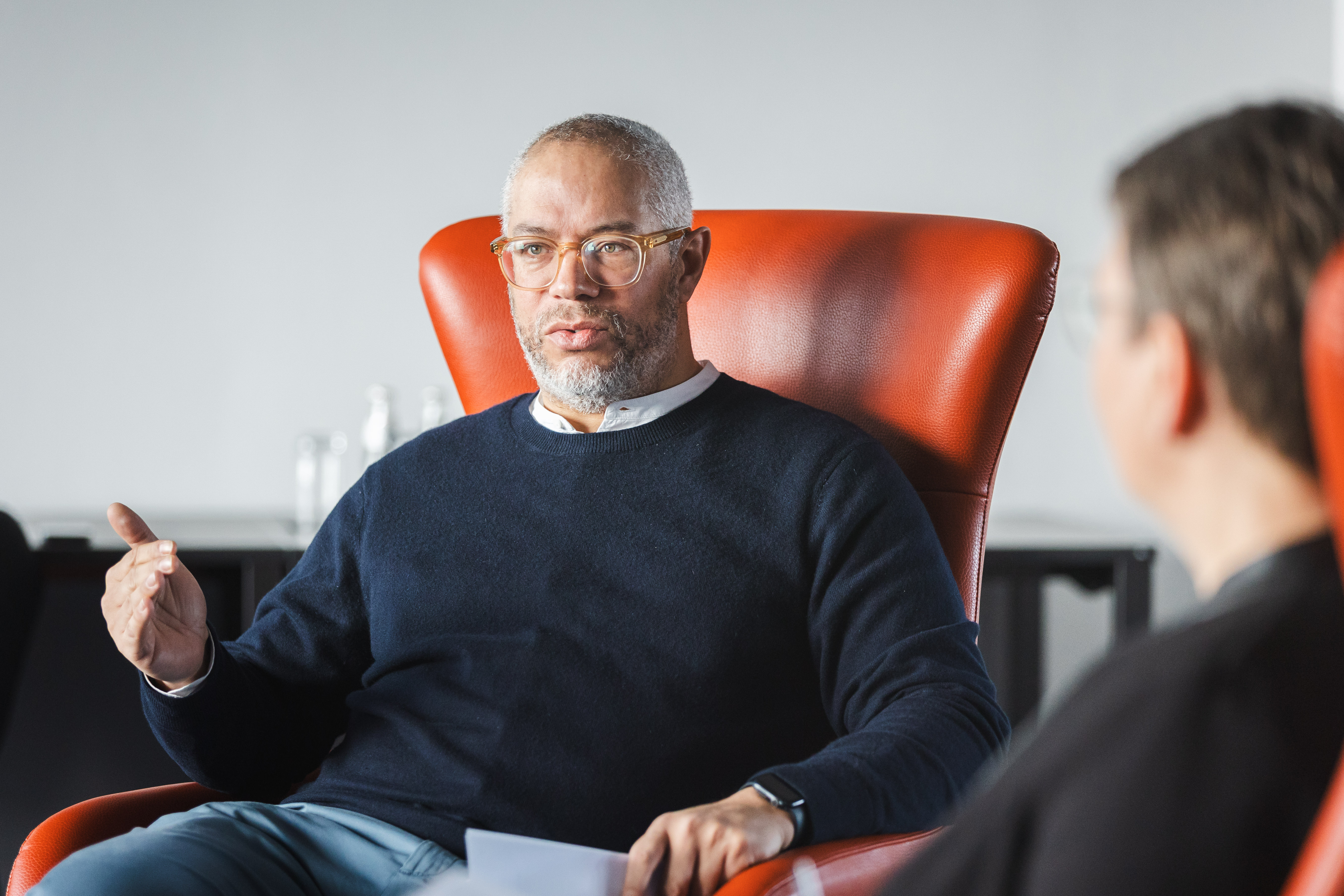 Porträtaufnahme eines Mannes mit Brille und grauem Bart, der in einem großen, orangefarbenen Sessel sitzt. Er trägt einen dunkelblauen Pullover und spricht konzentriert mit einer Person, die im rechten Vordergrund nur angeschnitten und unscharf zu sehen ist. Er unterstreicht seine Worte mit einer Handbewegung.