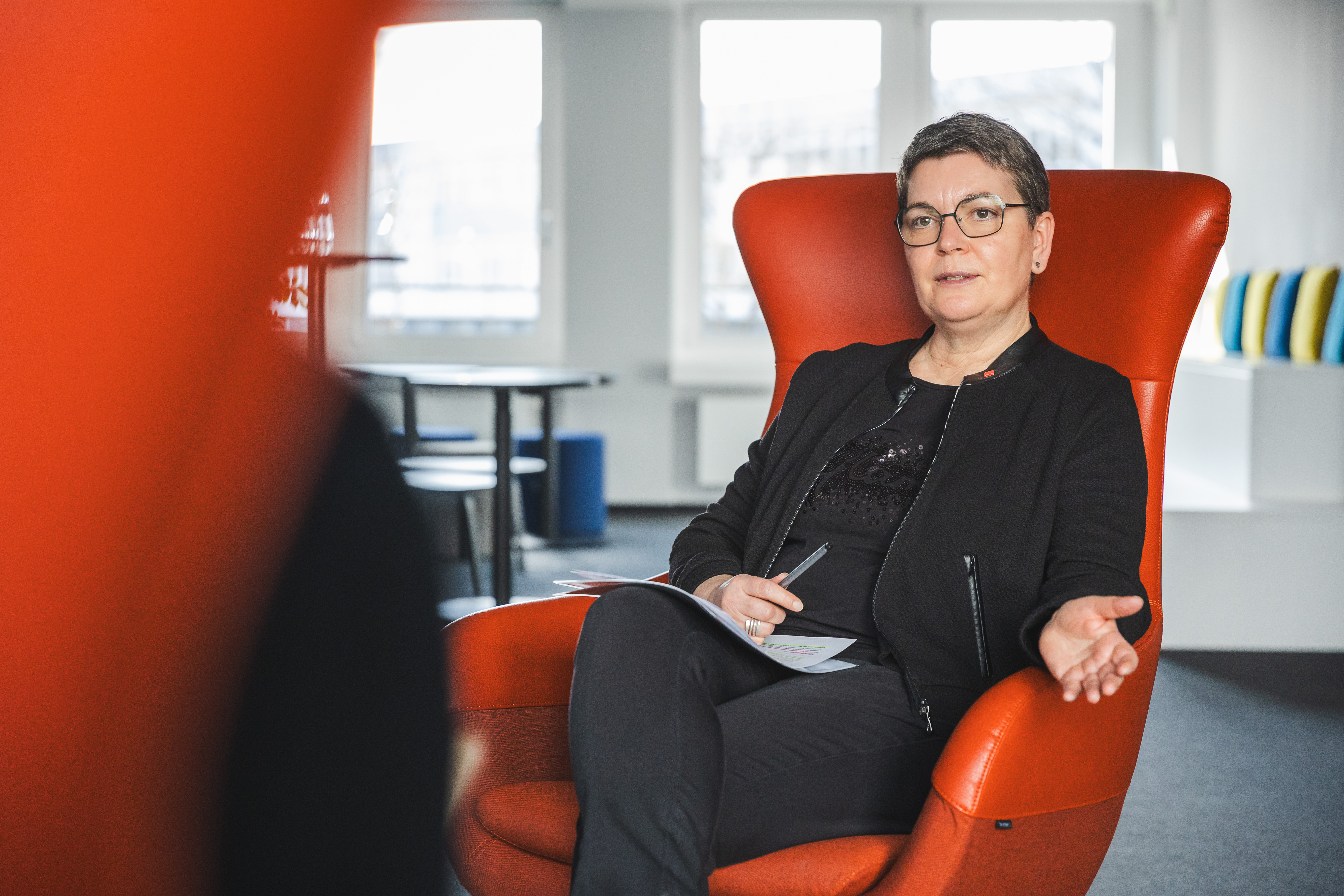 Zwei Personen sitzen sich in großen, orangefarbenen Ohrensesseln in einer modernen Büroumgebung gegenüber und führen ein Gespräch. Die Frau im Hintergrund trägt eine Brille und eine schwarze Jacke, hält einen Stift in der Hand und spricht gestikulierend. Der Mann im Vordergrund ist nur von hinten zu sehen. Auf einem kleinen Tisch neben ihnen stehen Wasserflaschen und ein Smartphone.