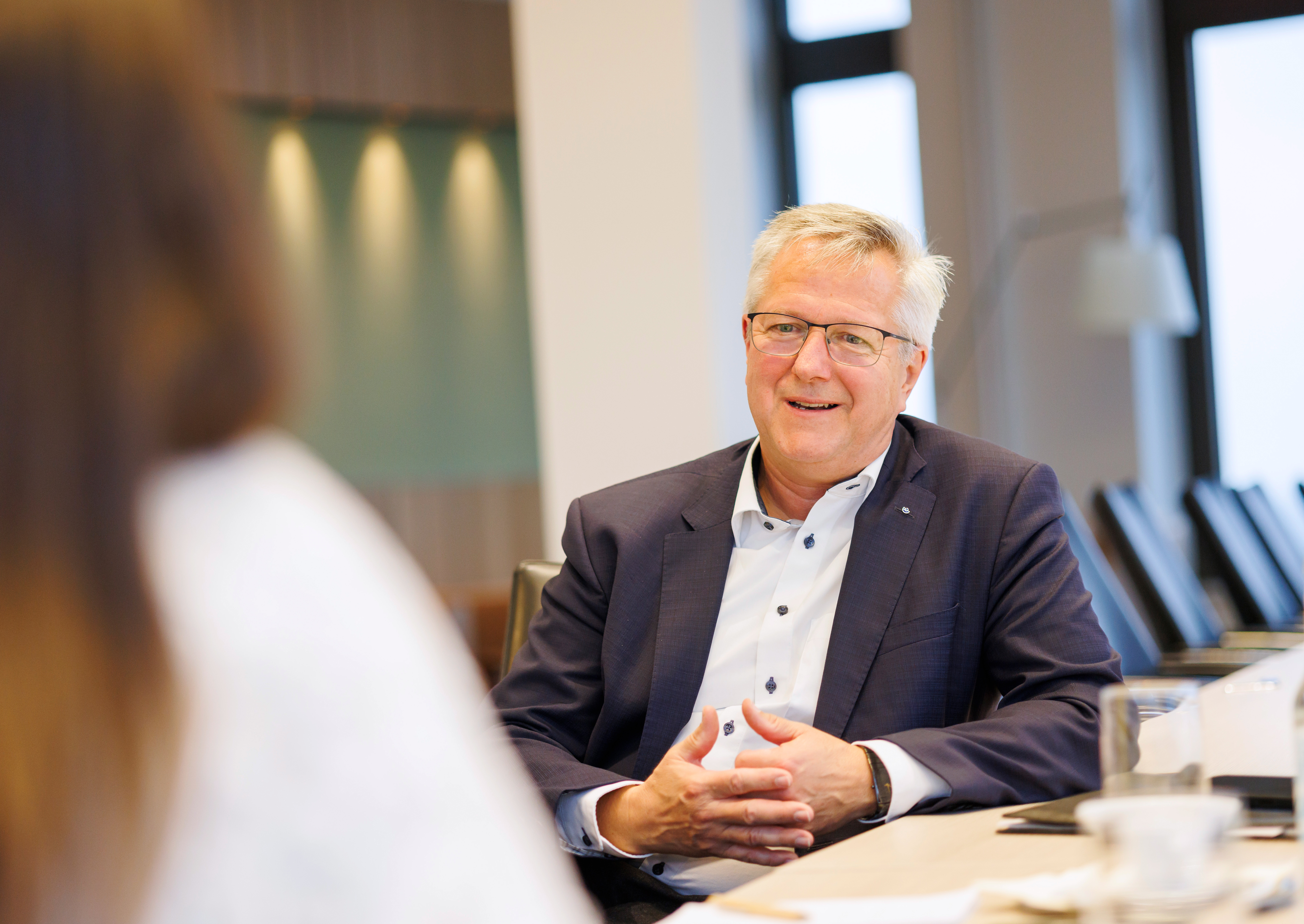 Ein Mann sitzt am Tisch. Man sieht nur den Überkörper. Er lehnt sich auf seinen linken Ellbogen. Er trägt ein weißes Hemd und ein dunkles Jackett. Er spricht und lächelt leicht. Er trägt eine Brille und hat graues Haar. 