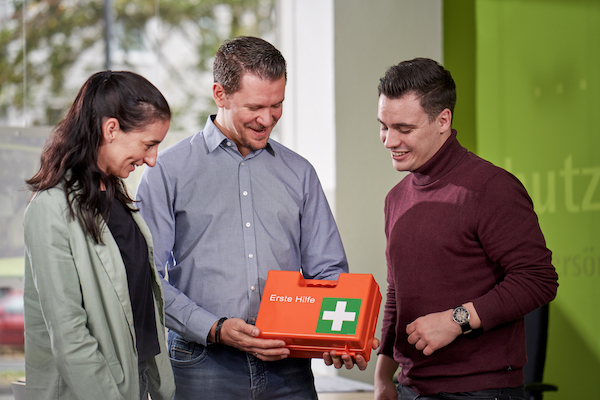 Foto: Zwei Männer und eine Frau stehen im Kreis und unterhalten sich. Sie lächeln. Einer der Männer präsentiert einen organgefarbenen Erste-Hilfe-Koffer.