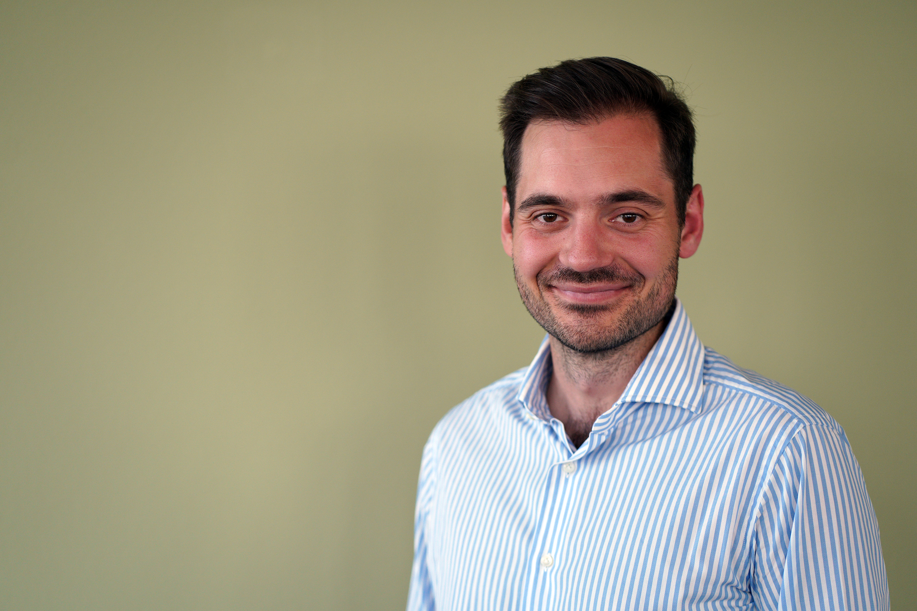 Ein Mann lächelt in die Kamera. Er trägt ein weiß, blaues Hemd. Er hat schwarze Haare  und einen leichten Bart.