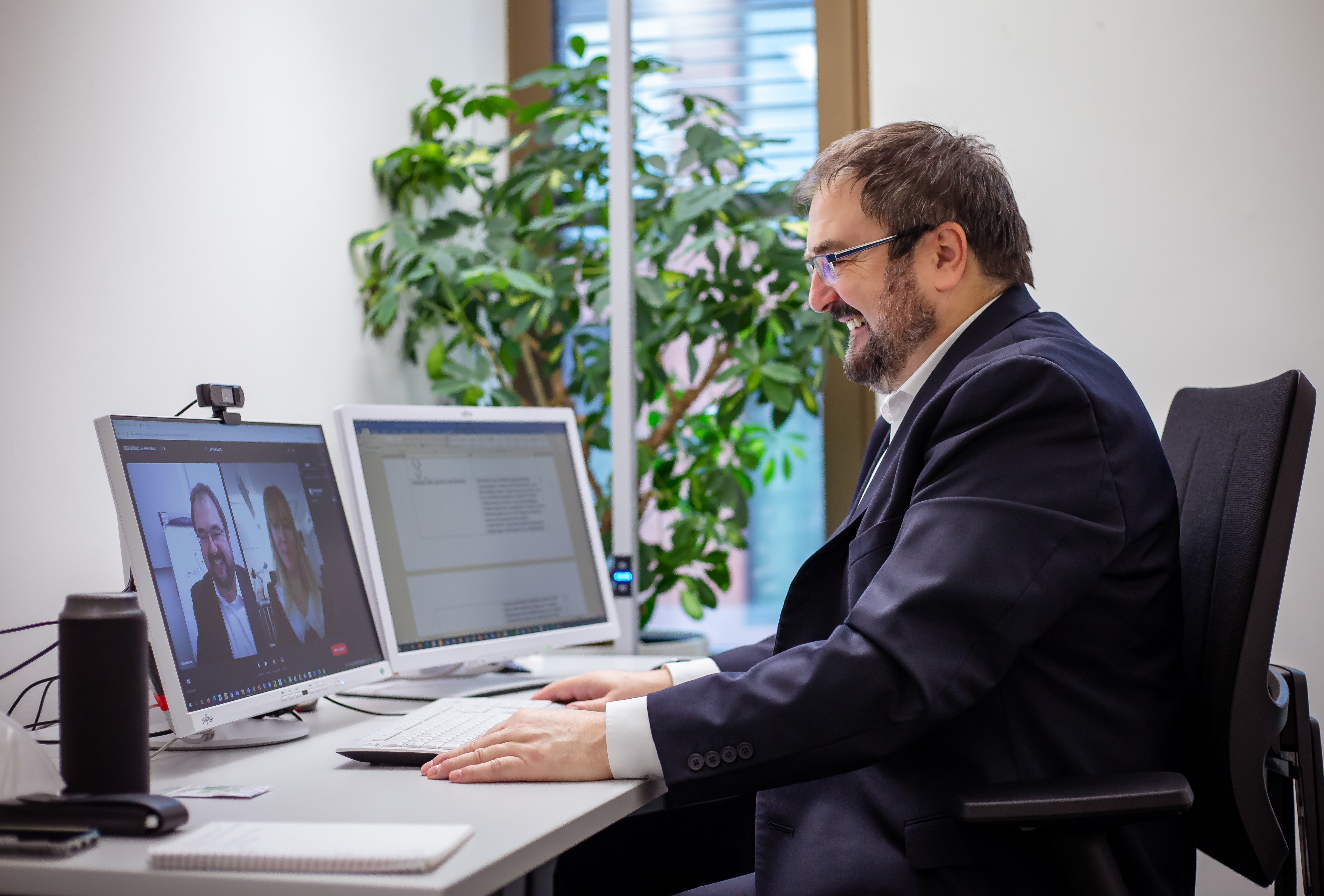 Ein Mann mit Brille, Bart und einem schwarzen Jacket sitzt an einem Schreibtisch. Man sieht eine Tastatur und einen Bildschirm. Der Mann ist in einem online-Meeting.