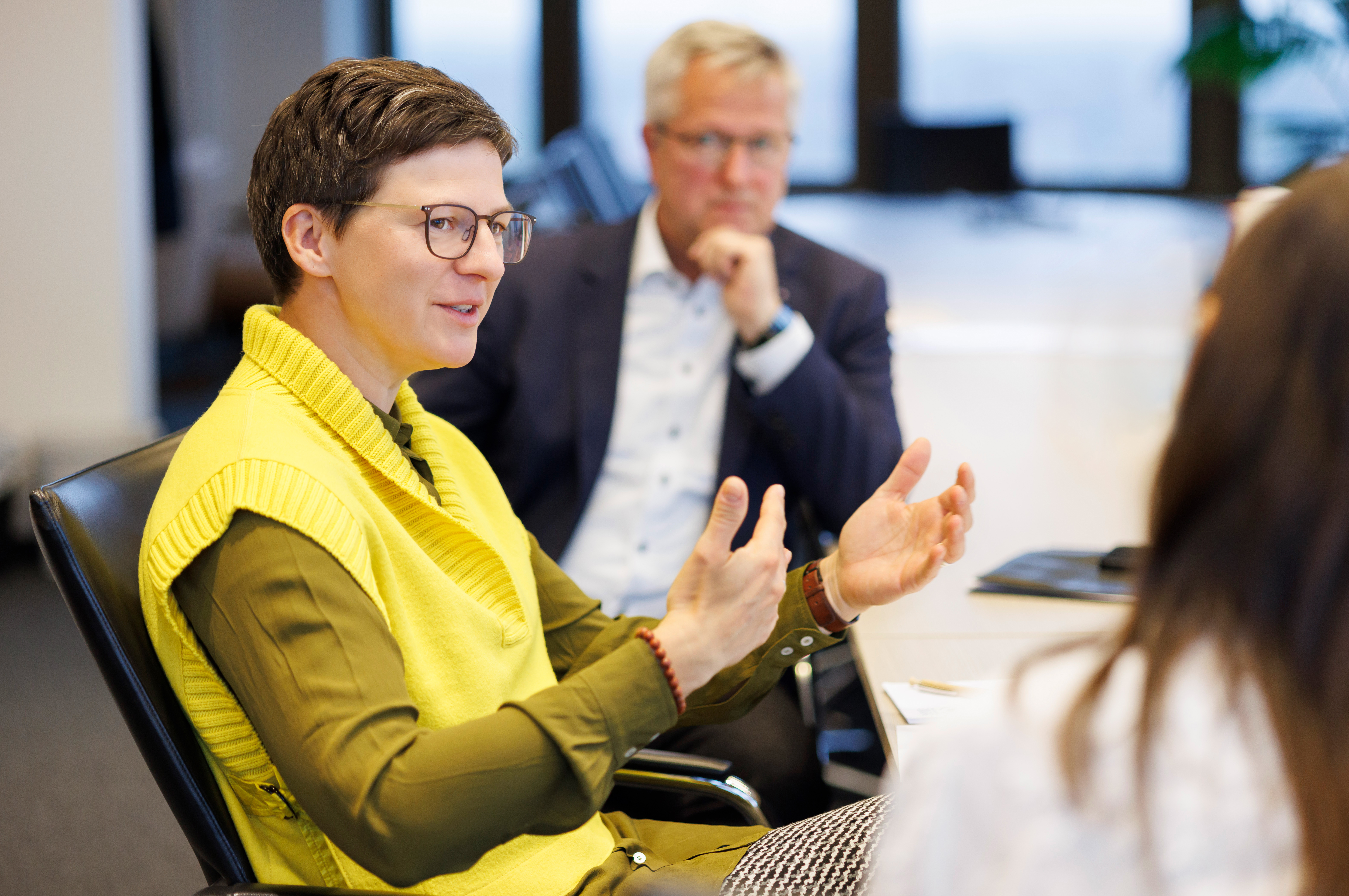 Eine Frau sitzt am Tisch und spricht. Man sieht sie von der Seite. Sie hat einen gelben Pullunder und eine grüne Bluse an. Sie hat schwarzes, kurzes Haar und eine dunkle Brille. Sie gestikuliert mit ihren Händen vor ihrem Körper und spricht.  