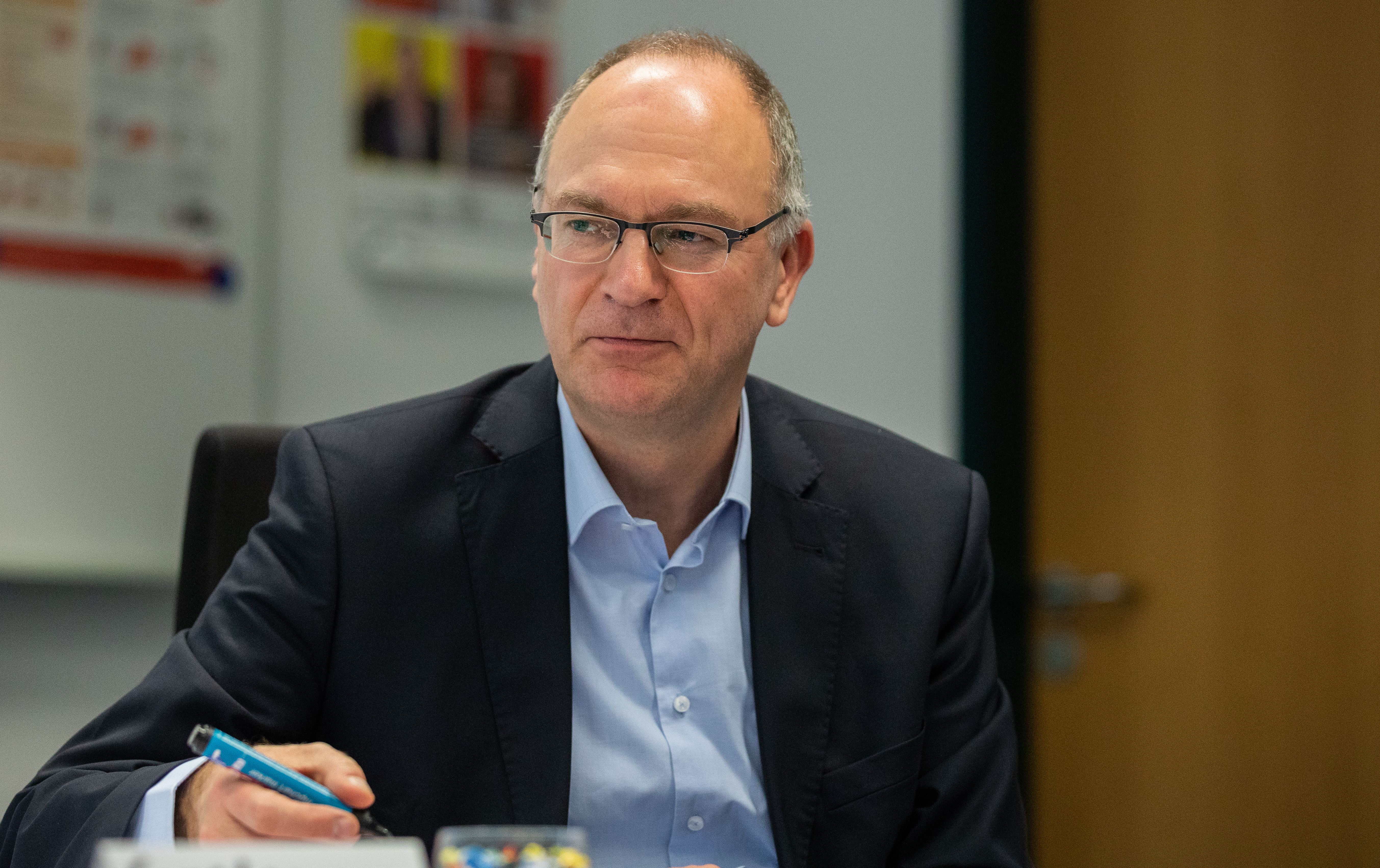 Porträt eines Brillenträgers in einem dunklen Sakko und einem hellblauen Hemd. Er sitzt an einem Tisch in einem Büro und blickt aufmerksam zur Seite. Im Hintergrund sind eine Tür sowie ein unscharfes Poster an der Wand zu erkennen.