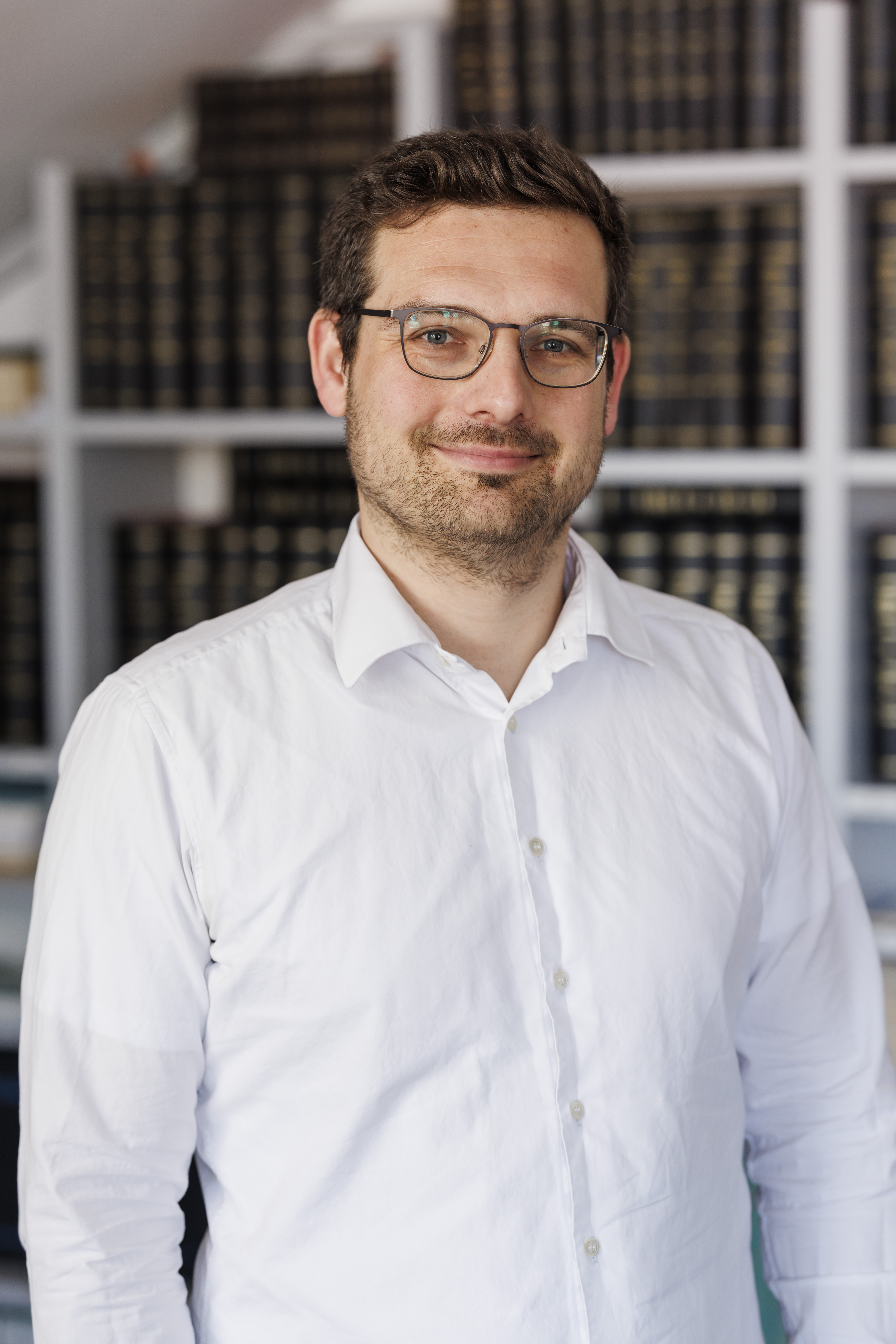 Ein Mann mit Brille und einem weißen Hemd steht lächelnd vor einem Bücherregal. Im Hintergrund sind viele Bücher zu sehen.