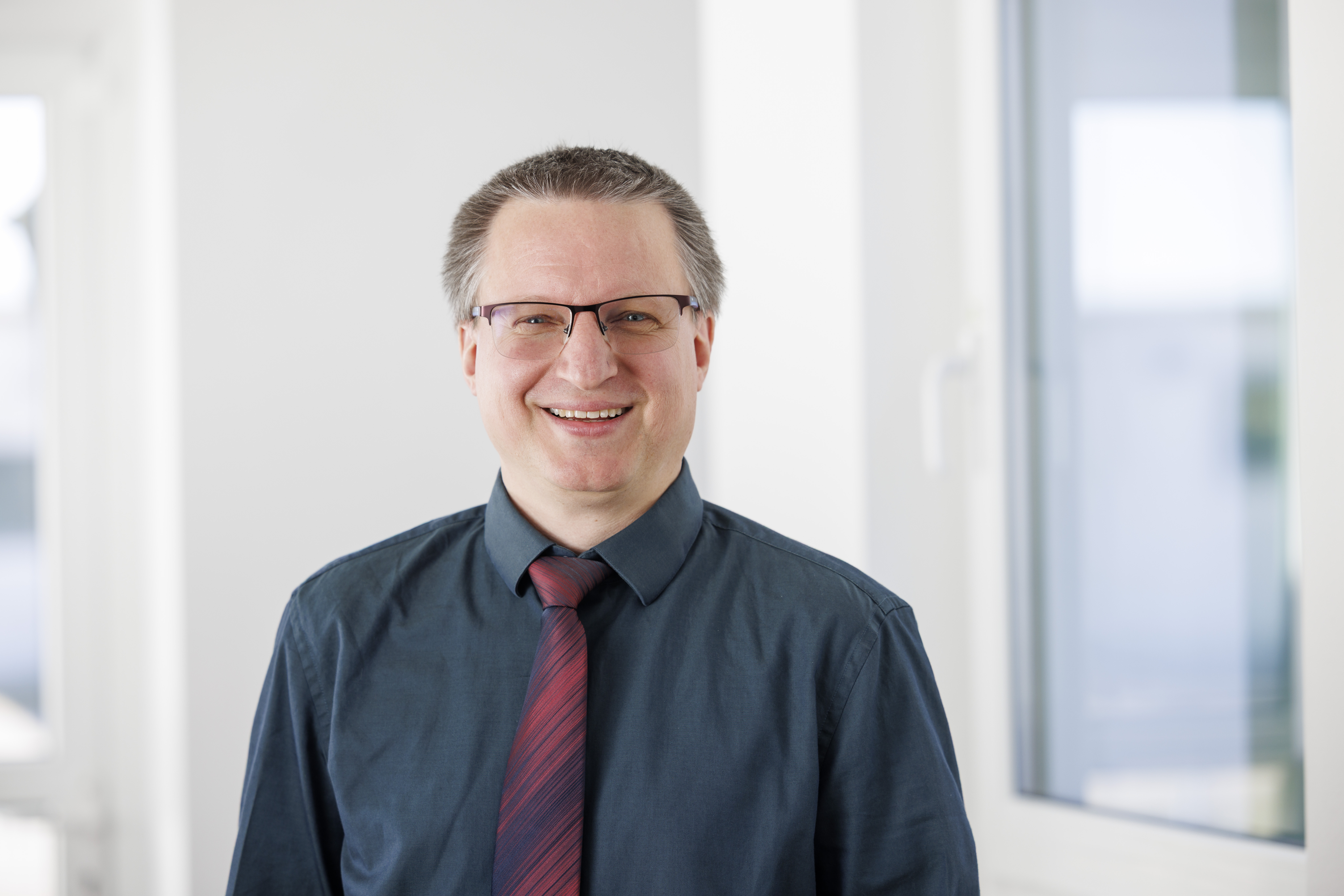 Ein Mann mit Brille und einem dunkelblauen Hemd steht lächelnd vor einem Fenster. Er trägt eine rot-schwarze Krawatte.