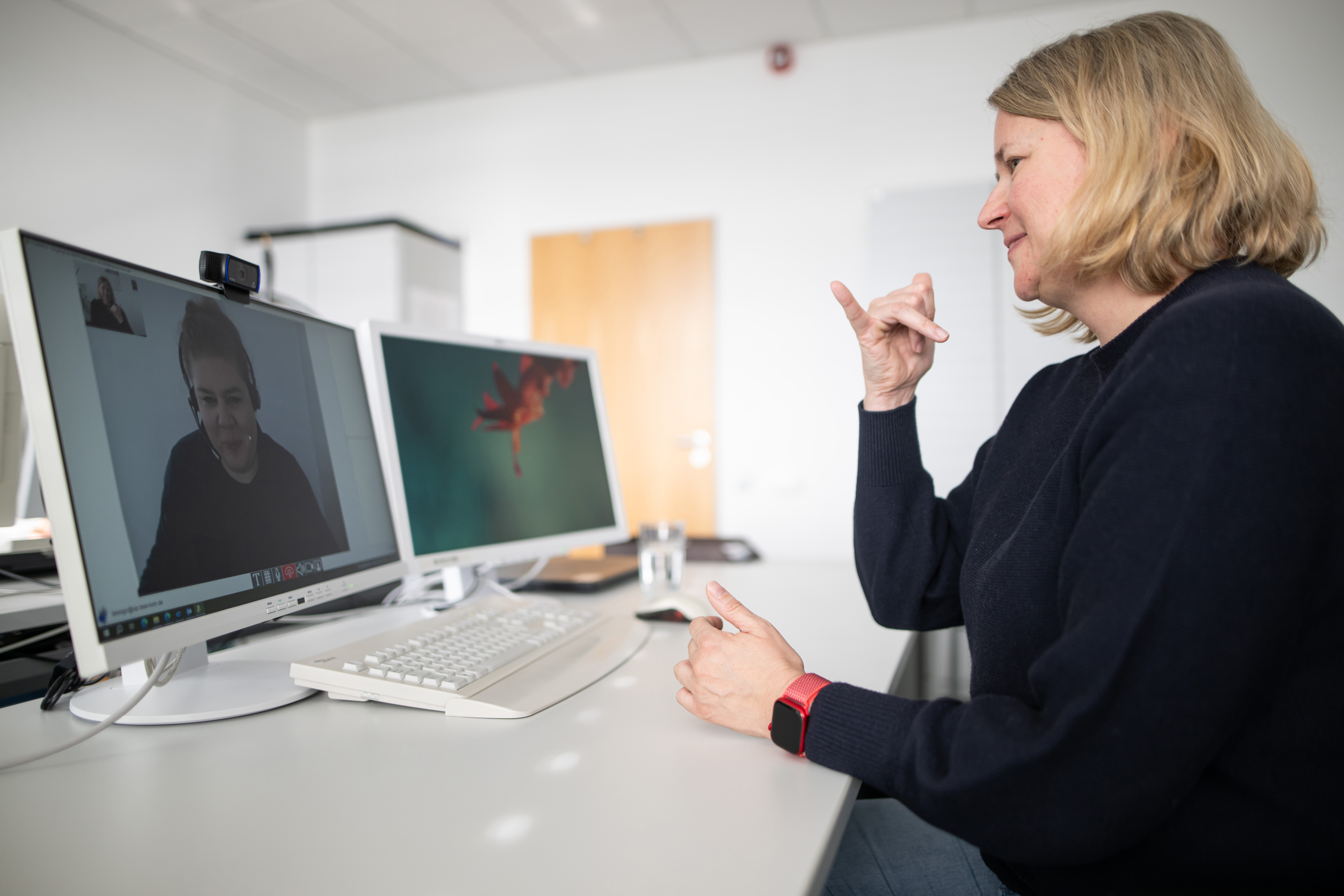 Foto: Sandra Fröhling sitzt mit dem Rücken zum Betrachter am Schreibtisch. Die Frau mach Gebärdensprache. Im Computerbildschirm auf dem Schreibtisch ist eine Gebärdendolmetscherin zu sehen. Die Dolmetscherin verfolgt die Gebärdensprache der Frau vor dem Bildschirm aufmerksam durch die Webcam.